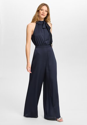 Femme portant une combinaison sans manches bleu marine avec un col montant orné d'un nœud et un pantalon à jambes larges, associée à des talons ouverts argentés.