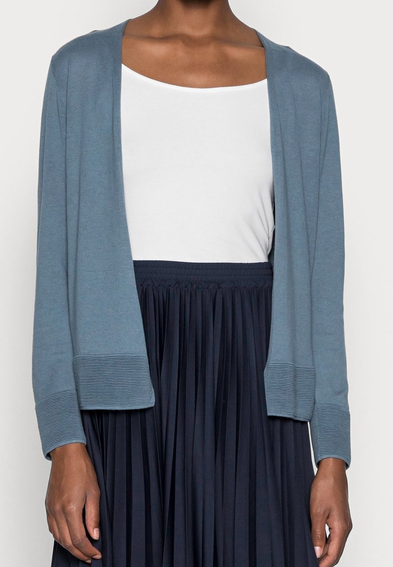 Hellblauer Strickcardigan mit entspannter Passform, offenem Vorderteil, gerippten Bündchen und Detail am Saum, über einem wei ßen Oberteil und einem marineblauen plissee Rock getragen.