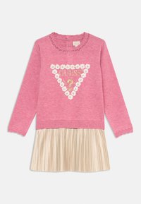Maglione rosa a maglia con scollatura arricciata e un design a triangolo che presenta "GUESS" e decorazioni floreali, abbinato a una gonna plissettata dorata.