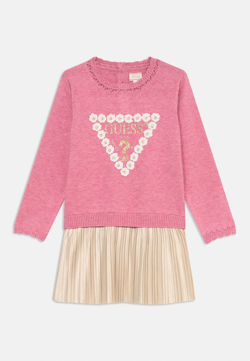 Maglione rosa a maglia con scollatura arricciata e un design a triangolo che presenta "GUESS" e decorazioni floreali, abbinato a una gonna plissettata dorata.