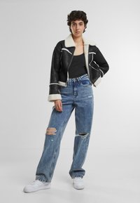 Veste noire cropped avec col et poignets en shearling blanc, associée à un jean léger bleu distendu et des baskets blanches.