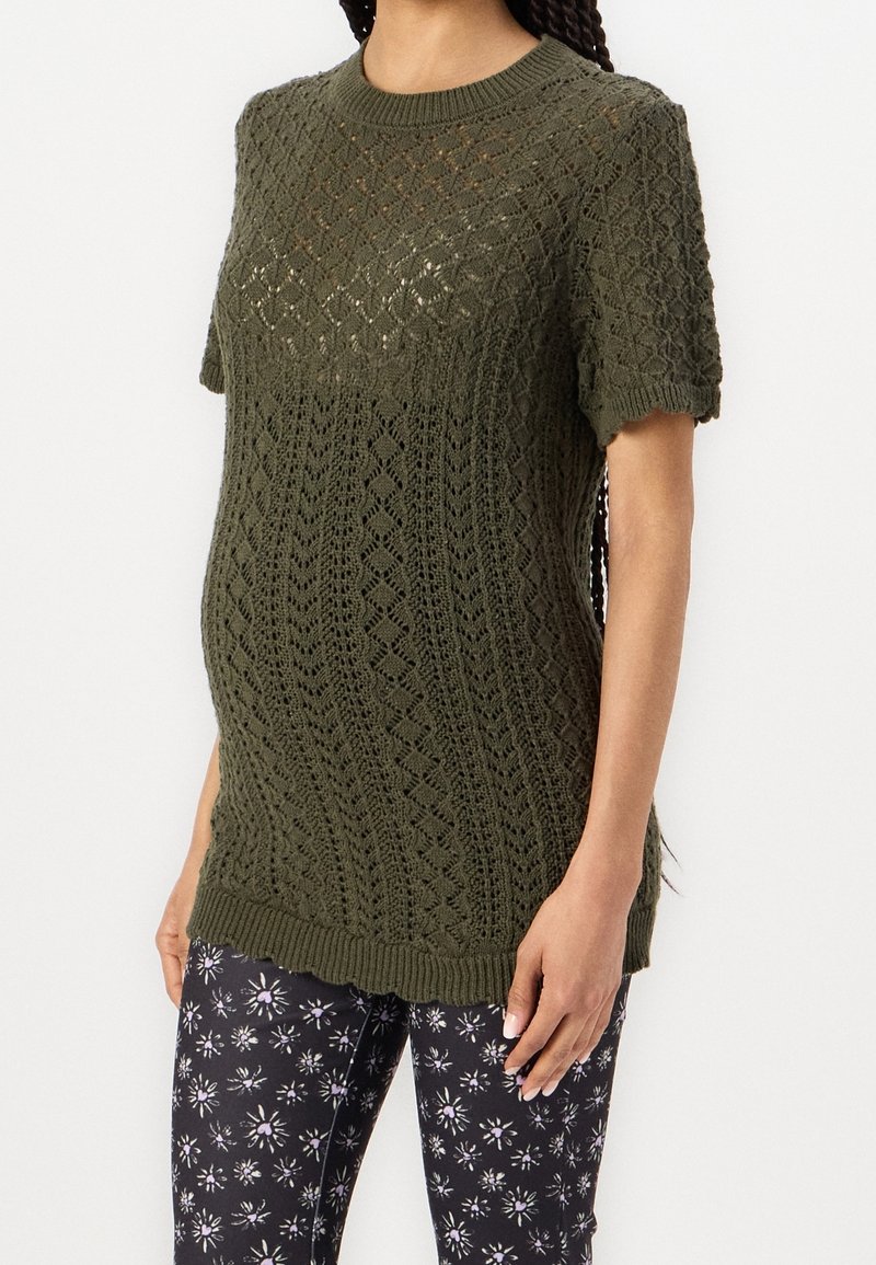 Frau in olivgrünem gestricktem Kurzarmpullover mit Rautenmuster und schwarzen Blumenleggings, stehend vor einfarbigem Hintergrund.