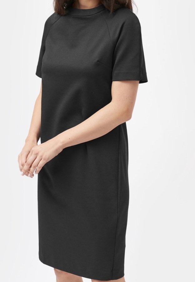 Femme portant une robe noire mi-longue à manches courtes avec un col montant, debout devant un fond blanc uni.