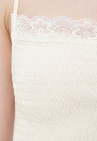 Gros plan d'un haut blanc en tissu élastique avec fines bretelles et une bordure en dentelle à motifs floraux au niveau de l'encolure.