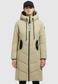 Gai bēšā krāsa pufīgs mētelis ar kapuci, zigzaga raksts, ar rāvējslēdzēju aizdari, melnas akcenti uz kabatām un vilkšanas auklām; valkāts ar melnu cepuri.