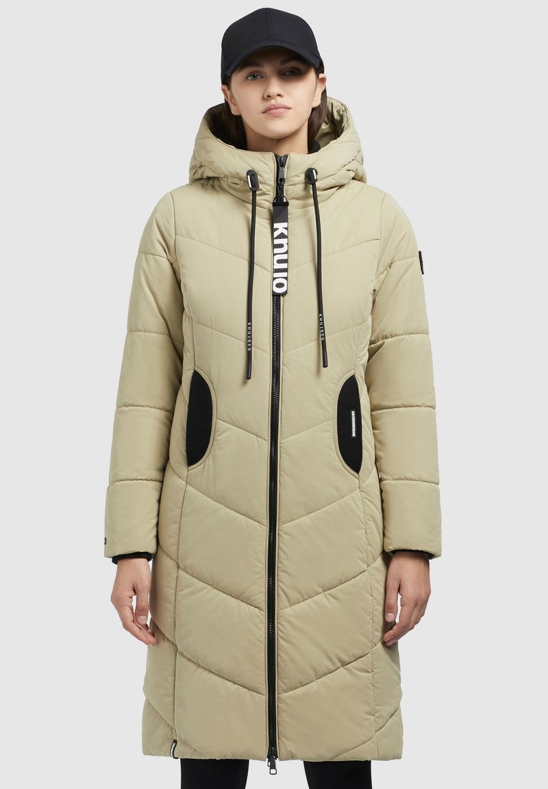 Gai bēšā krāsa pufīgs mētelis ar kapuci, zigzaga raksts, ar rāvējslēdzēju aizdari, melnas akcenti uz kabatām un vilkšanas auklām; valkāts ar melnu cepuri.