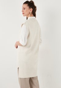 Lang creme strikket vest med sideslidser, båret over en hvid skjorte. Har en subtil tekstur og ribbet kant for ekstra detalje.