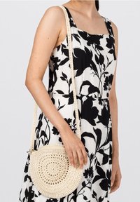 Rund vävd väska i off-white med spiraldesign. Har en lång, texturerad rem. Bärs som crossbody över en svartvit blommig klänning.