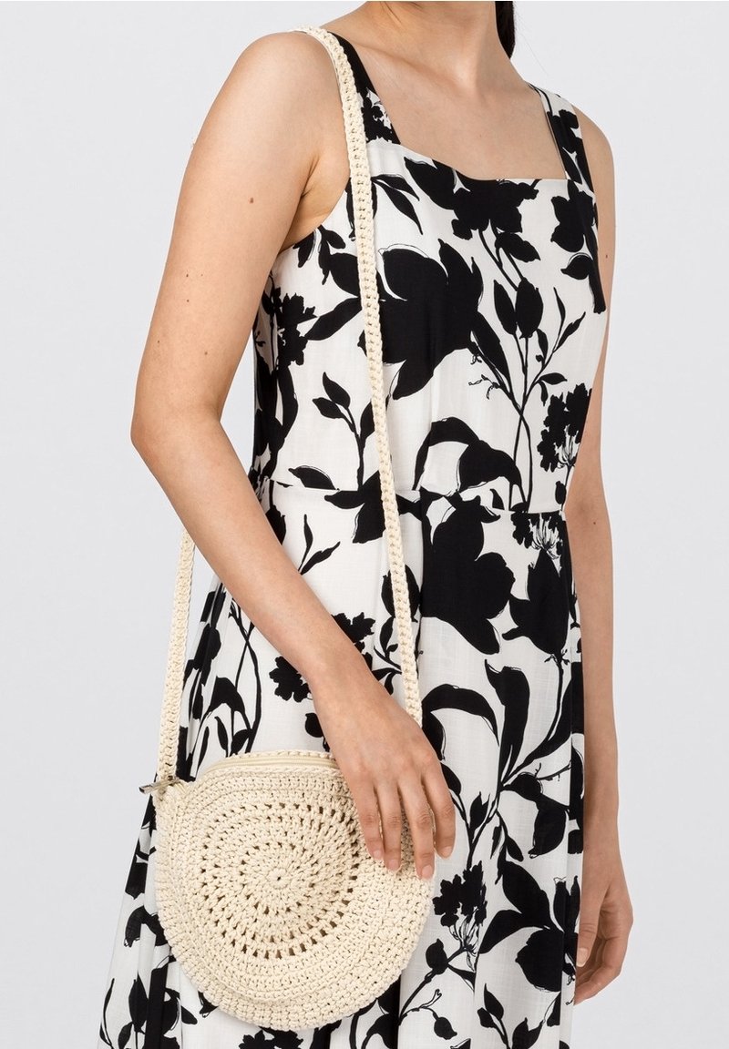 Rund vävd väska i off-white med spiraldesign. Har en lång, texturerad rem. Bärs som crossbody över en svartvit blommig klänning.