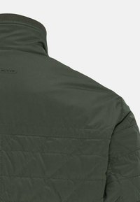 Mørkegrøn quiltet jakke med stående krave, tekstureret stof og diskret logo nær halsudskæringen. Har diagonale syninger.