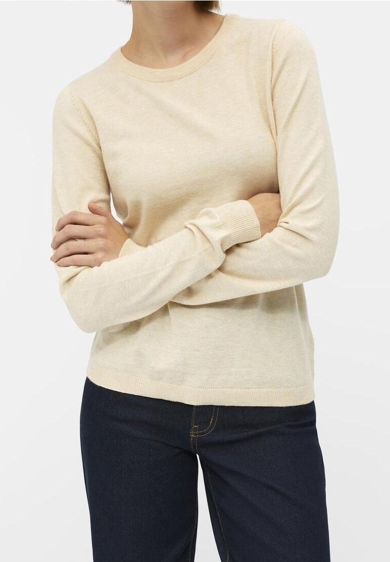 Femme portant un pull en maille beige à manches longues avec col rond et un jean bleu foncé, les bras croisés à la taille.