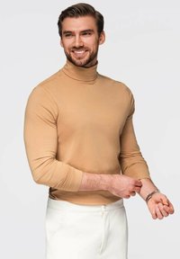 Hellbeige Rollkragenoberteil aus elastischem Stoff, mit langen Ärmeln, taillierte Passform, getragen zu weißen Hosen. Hände, die ein Armband am Handgelenk einstellen.