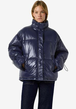 Femme portant une doudoune bleu foncé brillante avec un col montant et les mains dans les poches, associée à un jean foncé sur un fond uni.