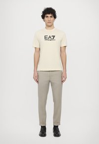Beige bavlněné tričko s černým logem EA7, spárované se světle šedými kalhotami a černými botami. Model stojí proti jednoduchému bílému pozadí.