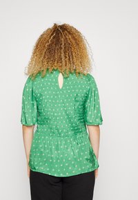 Grøn peplum-top med smocket ryg, korte puffærmer, blomsterprint og en nøglehulsdetalje ved halsen. Glat stofstruktur.