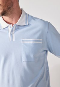 Ljusblå polo skjorta med vit krage, med en vänster bröstficka som har en vit horisontell rand. Tillverkad av strukturerad tyg.