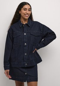 Temno modra denim jakna s srebrnimi konicami, velikimi žepnimi predelki in gumbi na pritisak. Ima sproščen kroj in manšete z gumbnimi dodatki.