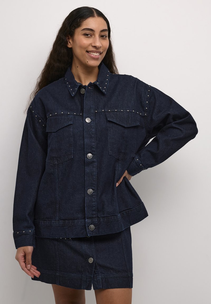 Temno modra denim jakna s srebrnimi konicami, velikimi žepnimi predelki in gumbi na pritisak. Ima sproščen kroj in manšete z gumbnimi dodatki.