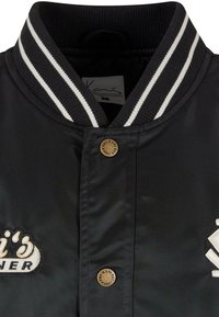 Fekete bomber dzseki bordás gallérral, fehér csíkokkal, hímzett foltokkal és bronz színű logógomb részlettel.