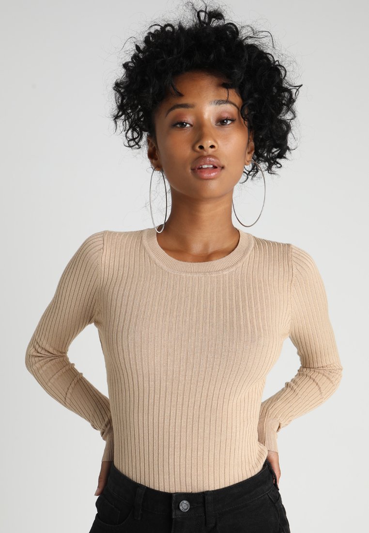 Haut beige à manches longues avec un design côtelé, une silhouette ajustée et un col rond, fabriqué à partir d'un matériau doux et extensible. Associé à un pantalon noir.
