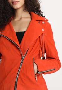 Veste bomber en daim rouge avec poches zippées, quincaillerie argentée et détails de franges sur les côtés. Présente un large col et une silhouette ajustée.