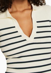 Gros plan sur une personne portant un pull côtelé crème avec des rayures horizontales noires et un col en V profond.