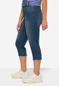Pantalones capri de mezclilla en lavado oscuro con un dobladillo ligeramente deshilachado, que cuentan con dos bolsillos traseros y una cinturilla ajustada. Combinados con zapatillas blancas.
