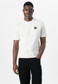 White short-sleeved t-shirt made of cotton, featuring a round neckline and a circular logo patch on the chest. Black jeans.