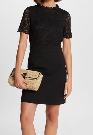 Black dress with lace short sleeves, scalloped neckline, and fitted waist. Holding a straw clutch bag with a black trim and clasp.