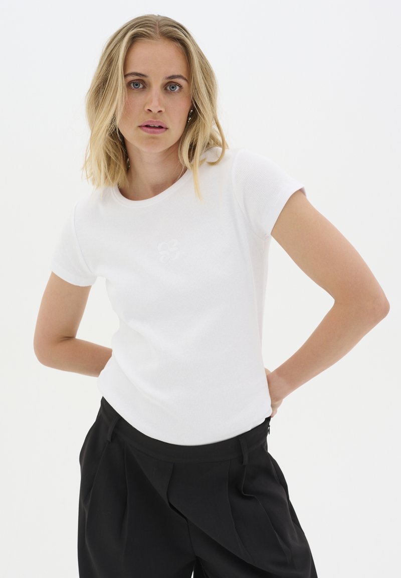 White short-sleeve t-shirt in ribbed fabric with a round neckline, featuring a subtle embroidered logo on the front. Paired with black trousers.