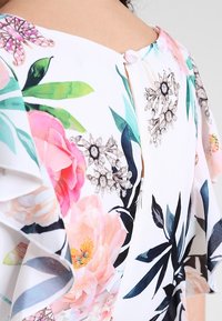 Chemisier à manches courtes en fleurs, avec une base blanche, des motifs de fleurs roses et vertes, et une fermeture à boutons dans le dos avec une fermeture éclair.