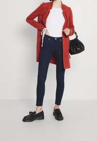 Cardigan lungo rosso, top aderente bianco, jeans skinny blu scuro con orlo sfrangiato, mocassini neri, borsa nera arricciata. Trame lisce.