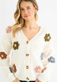 White knitted cardigan featuring large, textured flower appliqués in pink, blue, green, and orange. Four buttons down the front.