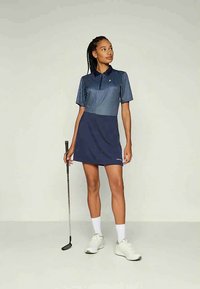 Sieviete zilās svītrām polo kreklā un svārkos, tur rokās golfa nūju, stāvot ar baltām sporta kurpēm un zeķēm uz vienkrāsaina fona.