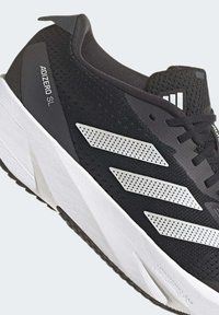 Black athletic shoe with a textured mesh upper and prominent white perforated three stripes. Features a thick white midsole and black outsole.