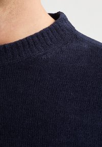 Pulover tricotat de culoare bleu marin cu guler rib. Materialul texturat are un aspect ușor păros și un croi care se mulează pe gât.