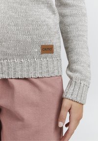 Maglione a maglia grigio chiaro con orlo e polsini a coste. Presenta una piccola toppa marrone con il logo "oxmo". Abbinato a un capo rosa.