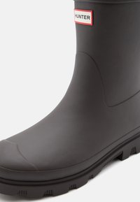 Tmavě šedá gumová holínka s kulatou špičkou, texturovaným povrchem a vyznačenou nášivkou loga s červenými a bílými akcenty.