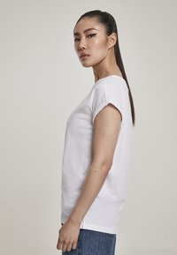 Balts crew-neck T-krekliņš ar īsām rullētām piedurknēm, izgatavots no mīkstas kokvilnas, valkāts kopā ar džinsu biksēm, attēlots sānskatā.