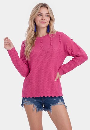 Kvinde med langt blond hår iført en lyserød tekstureret sweater og blå duskeøreringe, kombineret med slidte denimshorts, ser til siden.