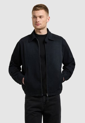 Jaunas vyras dėvi juodą užsegamą striukę ir juodą marškinėlį, stovintis rankomis kišenėse, žvelgiantis į kairę, paprastame pilkame fone.