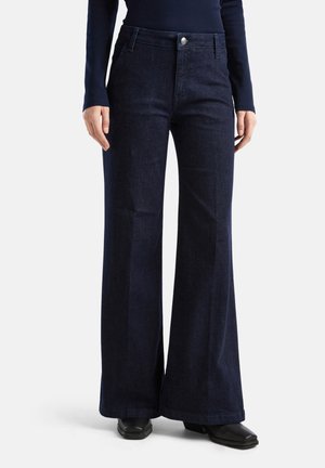 Jean bleu foncé taille haute à jambes larges porté avec des chaussures noires à bout carré et un haut bleu marine à manches longues, montré de la taille aux pieds.