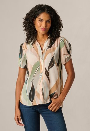 Vrouw met krullend haar die een kortemouws, knoopsluitende blouse draagt met abstracte beige, groene en zwarte bladpatronen, gecombineerd met een blauwe spijkerbroek.