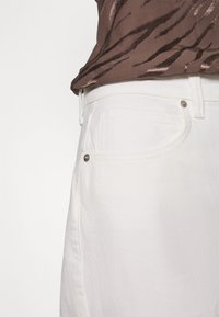 Calças de ganga brancas com um corte clássico, apresentando bolsos frontais e detalhes em tom prateado. Exibidas com uma blusa castanha estampada.