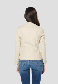 Licht beige leren jas met gewatteerd schouderdetail, aansluitend ontwerp en lange mouwen. Gecombineerd met blauwe spijkerbroek.