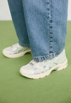 Osoba ubraną w jasnoniebieskie dżinsy i białozielone buty do biegania stoi na zielonej powierzchni.