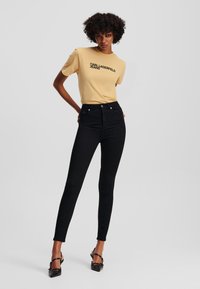 Camiseta amarilla de manga corta con el texto negro "KARL LAGERFELD JEANS", combinada con jeans ajustados negros de cintura alta y zapatos de tacón negros.
