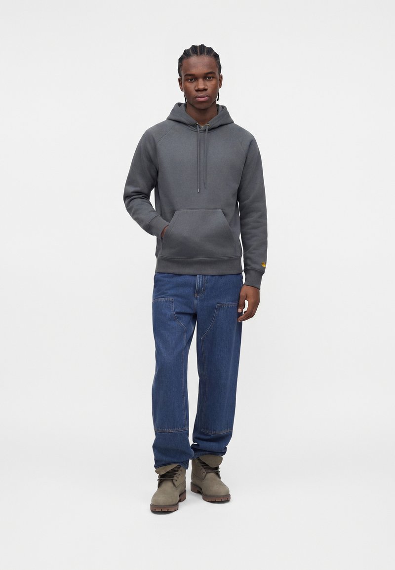 Mann steht und trägt einen grauen Kapuzenpullover, blaue Jeans mit aufgesetzten Taschen und beige Schnürstiefel vor einem schlichten weißen Hintergrund.