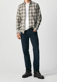 Camicia a quadri nei toni beige, blu e giallo; t-shirt bianca a girocollo sotto; jeans blu scuro; scarpe nere con suola texturizzata.