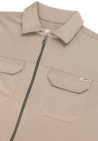 Chaqueta beige con cremallera y cuello puntiagudo, dos bolsillos delanteros con solapa y una pequeña etiqueta en el bolsillo derecho.
